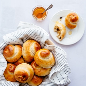 Sweet Challah rolls in a basket with a striped napkin and a plate with a challah roll on the side plus a glass of honey.