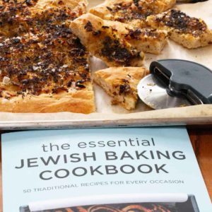 A pletzel on a parchment-lined tray with a pizza cutter with a copy of the Essential Jewish Baking Cookbook in front of the tray.
