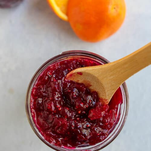 Cranberry orange jam straight down view in a jar with a wooden spoon inside and an orange in the background.