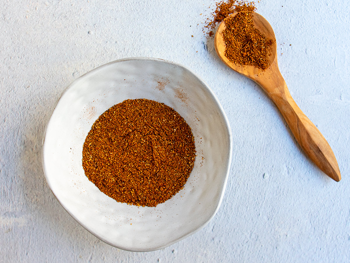 Baharat spice blend in a grey bowl with a wooden spoon holding some spice rub next to it.