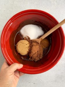 Sugars and cocoa added to melted butter and chocolate in red bowl.