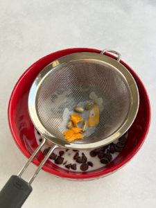 Strainer over red bowl with cardamom pods and orange peel in it.