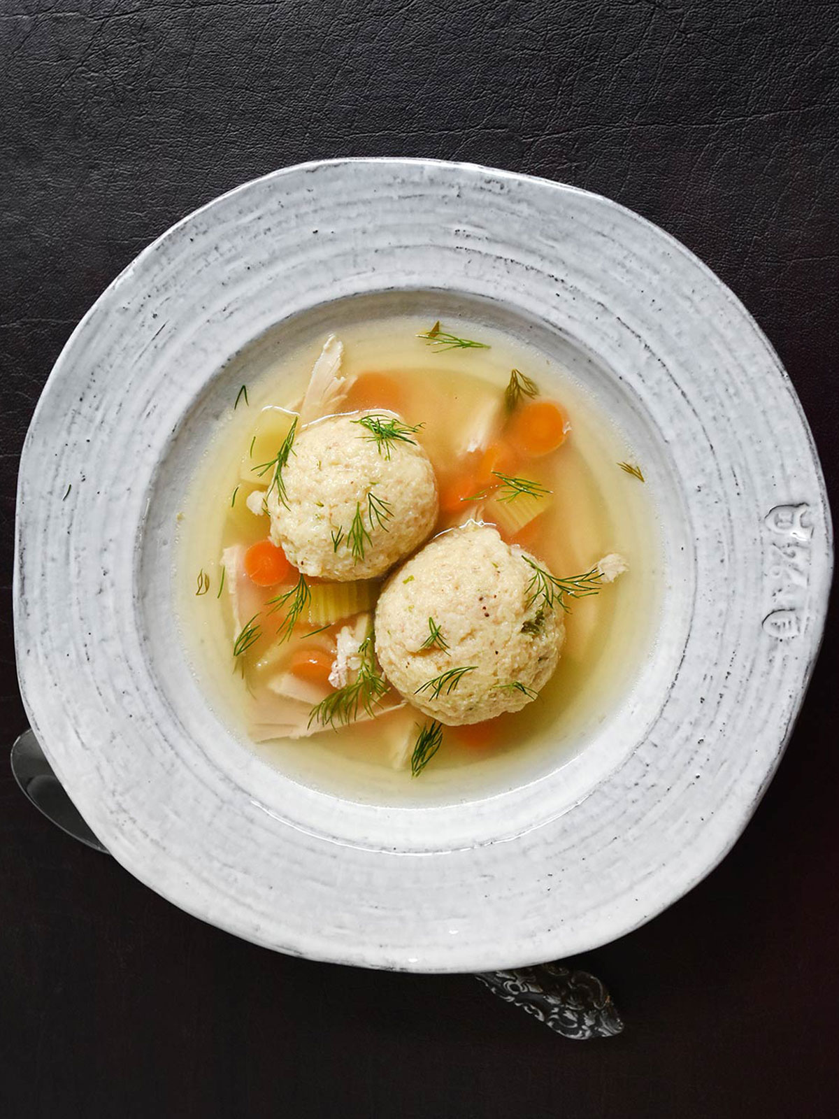 White bowl of matzo ball soup on a dark background.