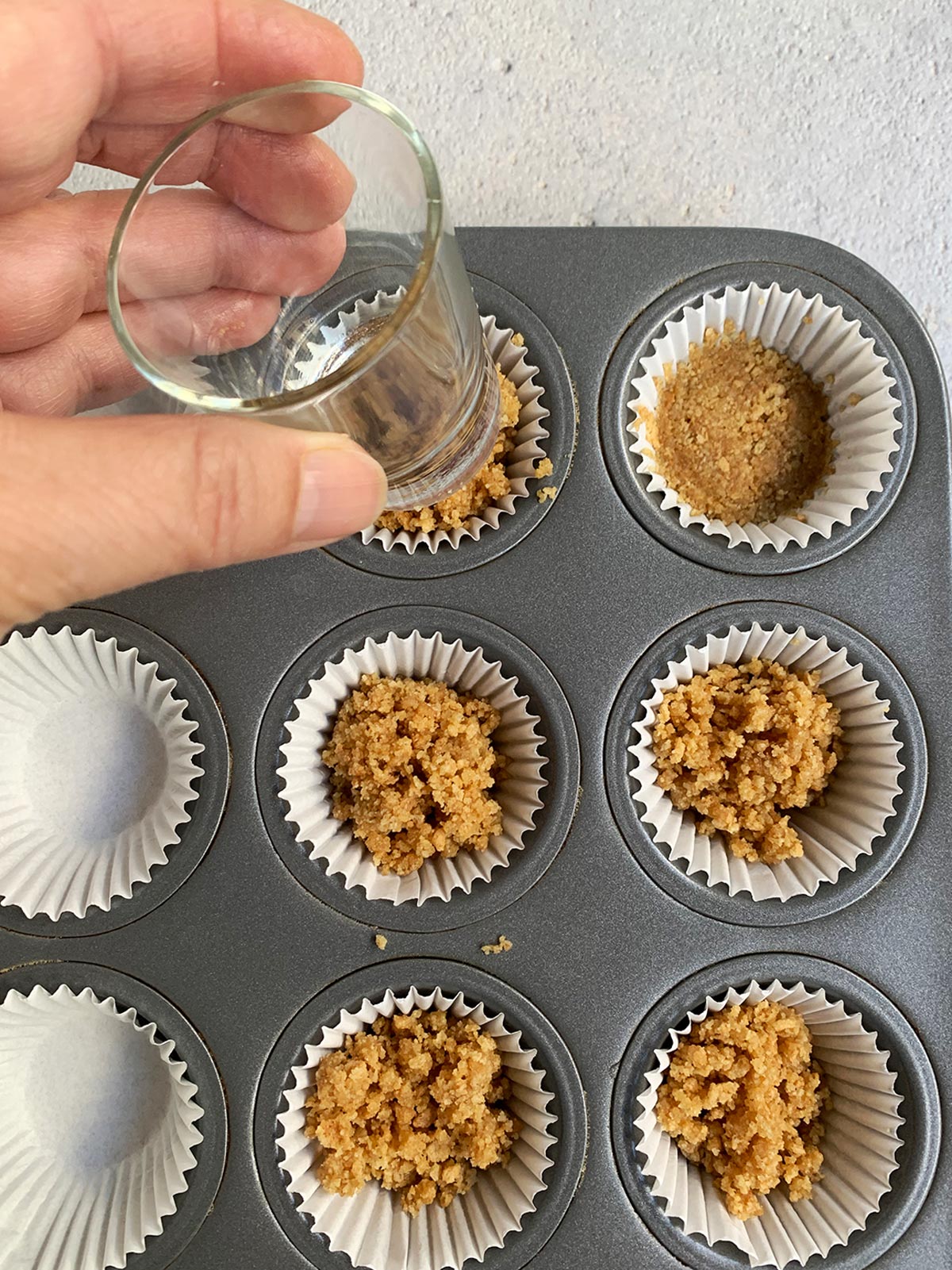 Graham cracker crust being pressed into mini muffin liner with a shot glass.