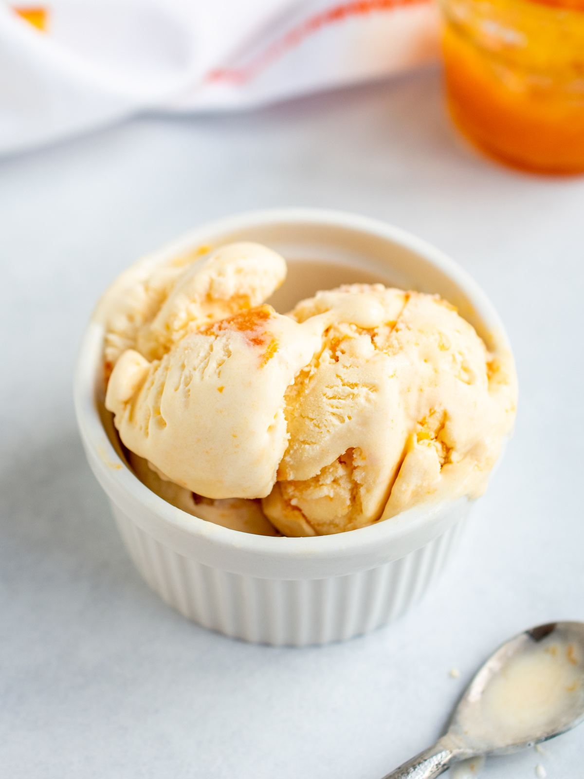 Scoop of apricot ice cream in white ramekin with silver spoon and jam jar in background.
