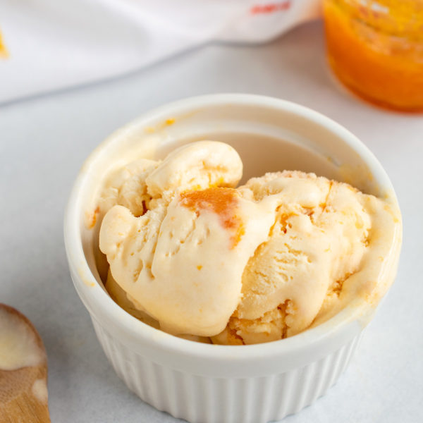 Close up angle of apricot ice cream in a white ramekin with a wooden spoon and jam in the background.