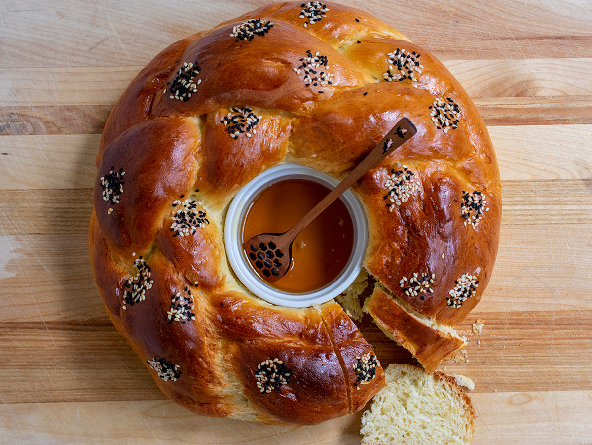 Round challah with a white ramekin in the center filled with honey and one slice cut out of the bread.