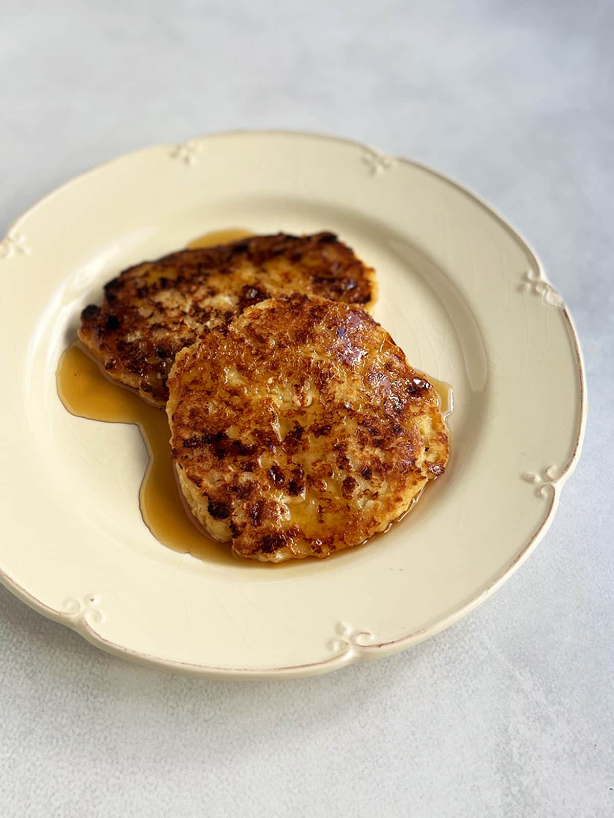 Two cheese latkes on an off white plate with maple syrup poured on top.