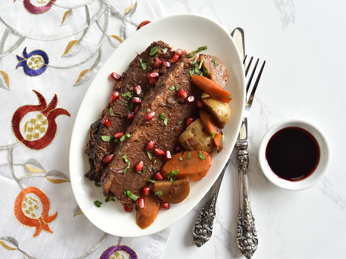 Slices of pressure cooker brisket with pomegranate on a white plate with potatoes and carrots sitting atop a pomegranate tablecloth.