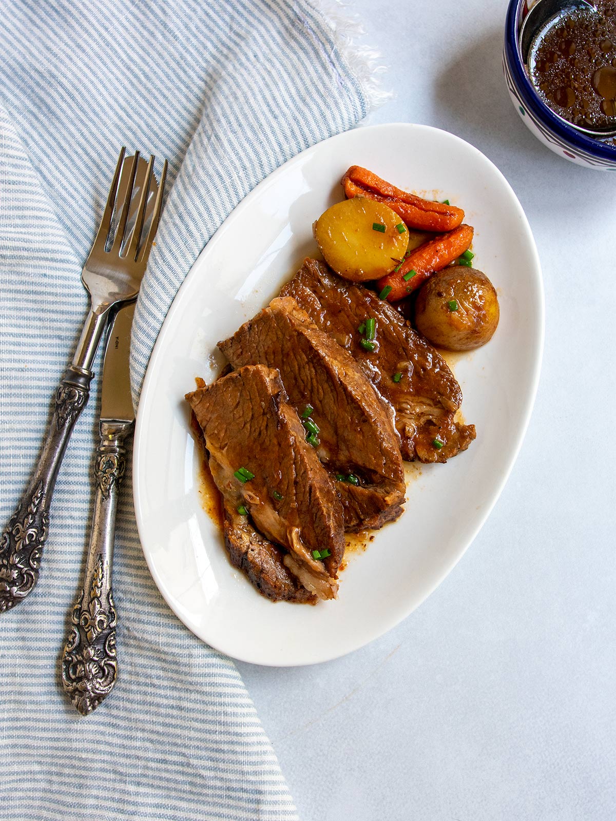 Sliced Jewish brisket on a small white plate with potatoes and carrots and knife and fork nearby.