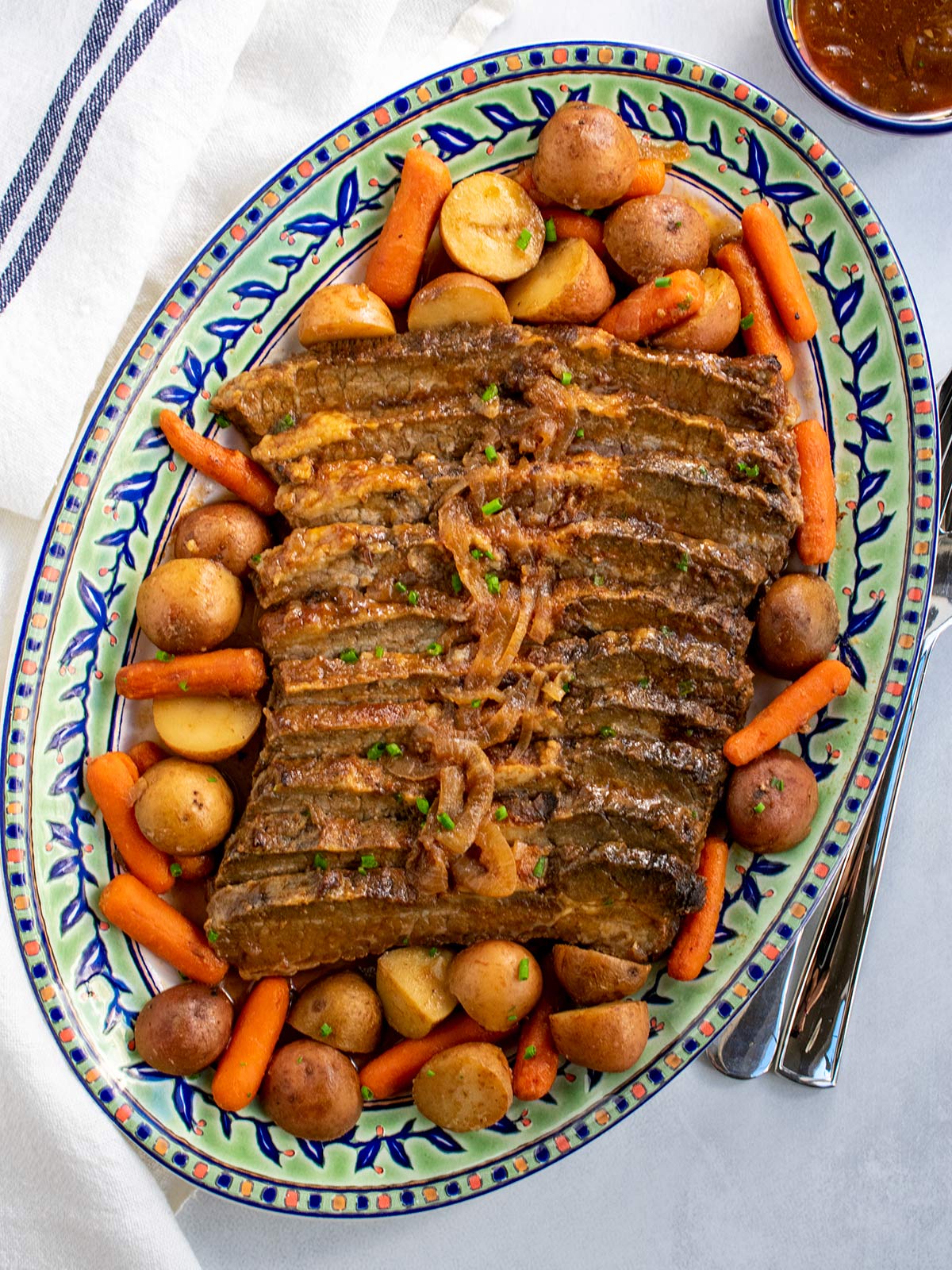 A platter of brisket with potatoes and carrots surrounding the meat and some gravy in a small bowl on the side to introduce the brisket section of a post on Rosh Hashanah food ideas.