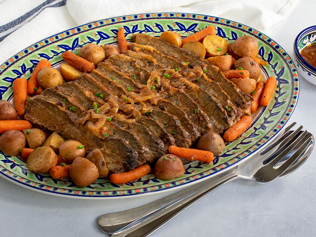 Angled view of large platter of Jewish brisket with gravy on the side and serving utensils.