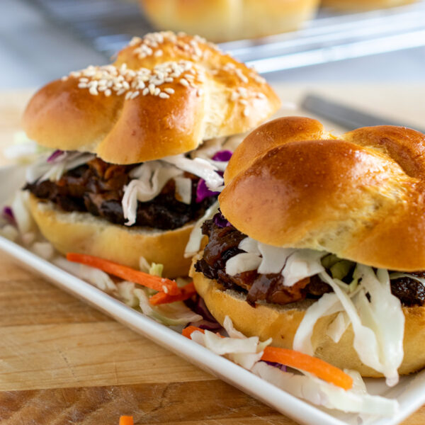 Two brisket sandwiches on a white plate with a cooling rack of slider rolls in the background.