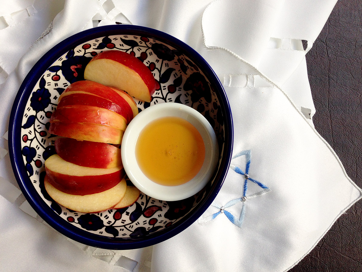 Top down view of a blue bowl with sliced apples and honey in it on a Jewish designed table runner.