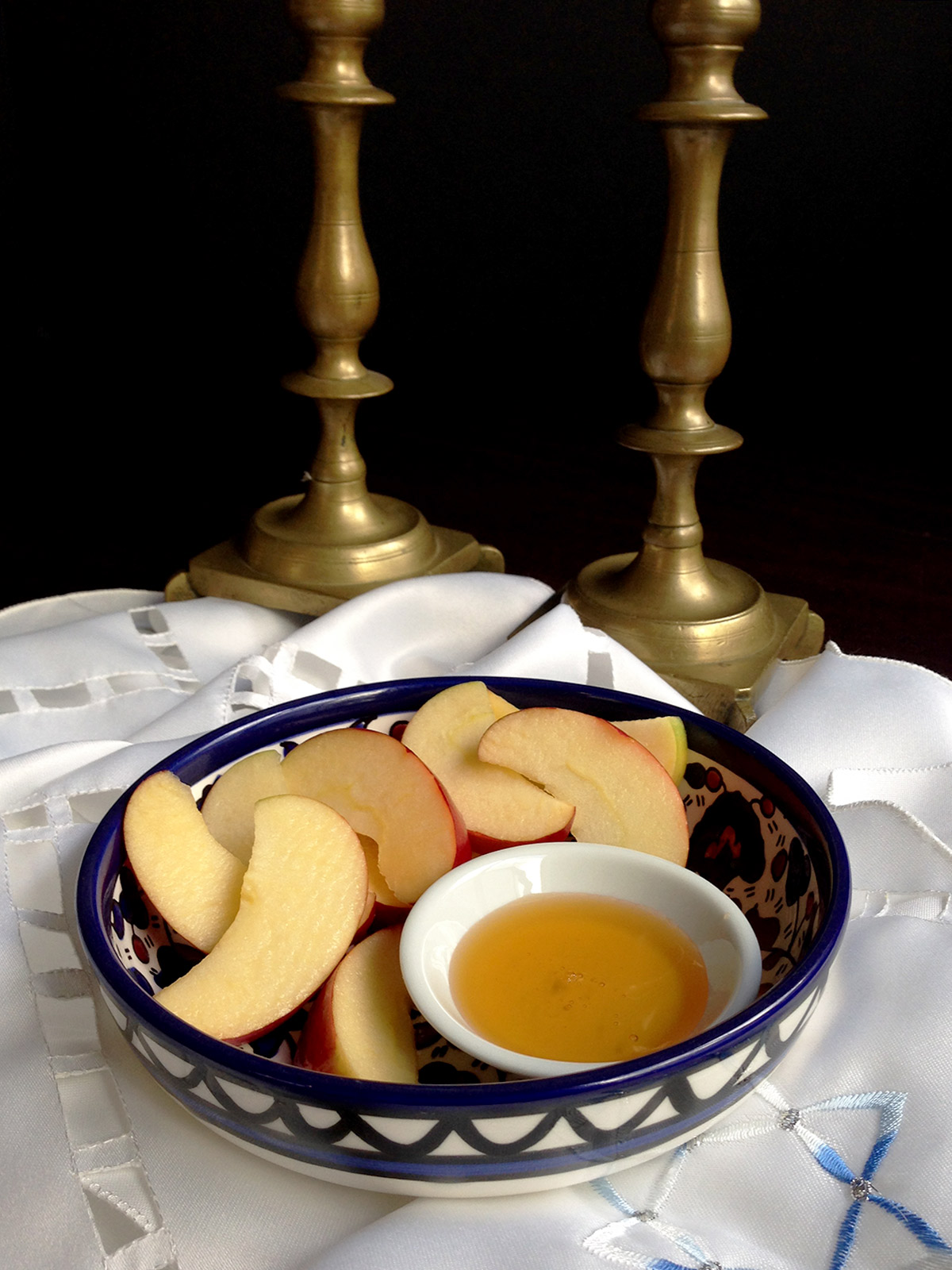 Apples and honey in a blue bowl with two shabbat candlesticks in the background.