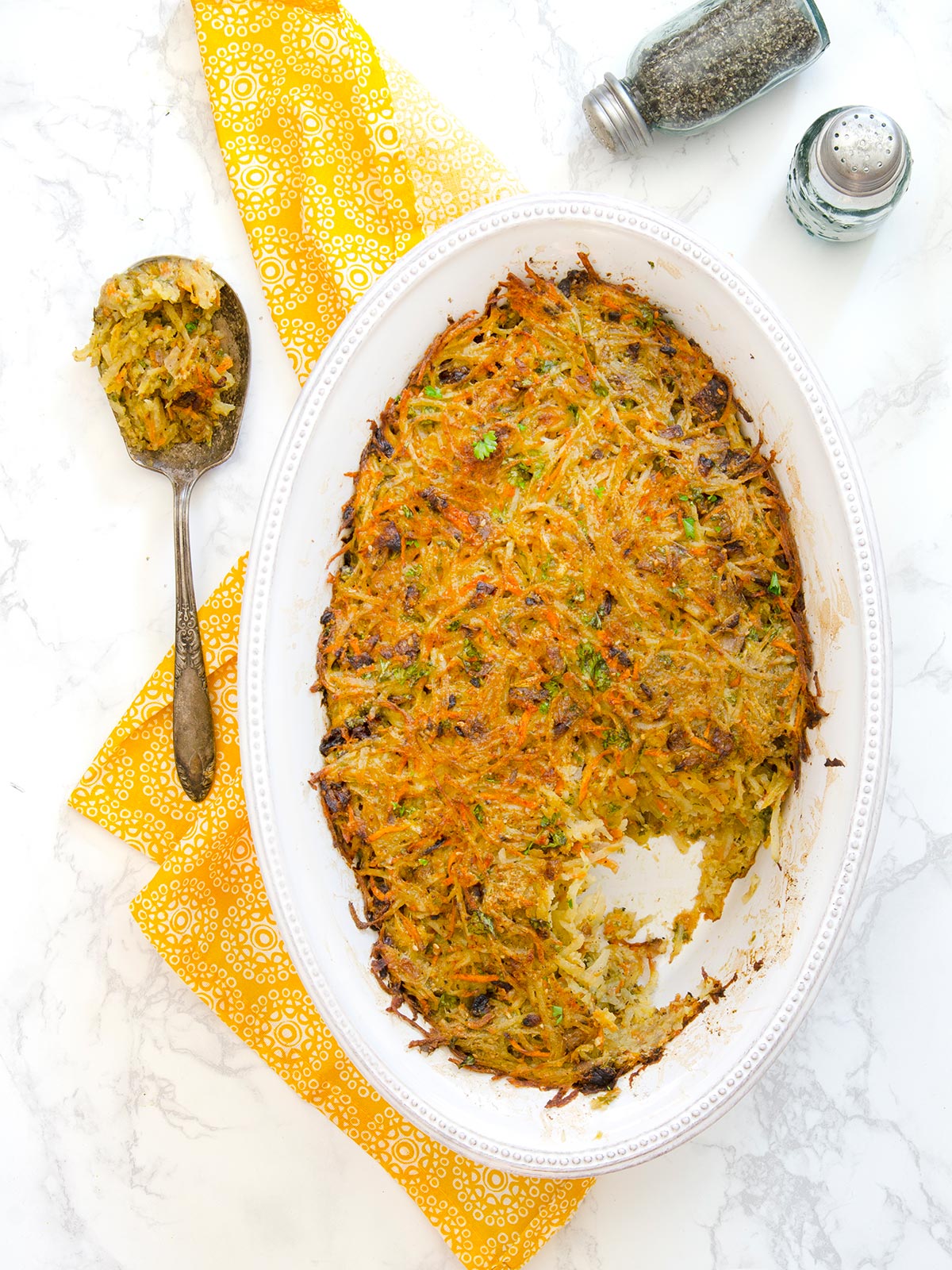 Top down view of a whole oval white pan of potato kugel with a spoon of it on the side and some salt and pepper shakers.