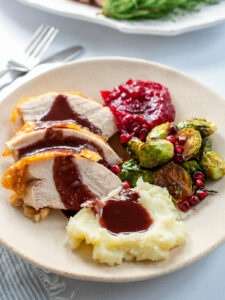 Sliced turkey, mashed potatoes, pomegranate gravy, cranberry jam and brussels sprouts on an off white plate.