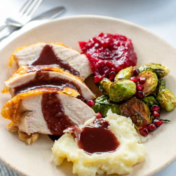 Sliced turkey, mashed potatoes, pomegranate gravy, cranberry jam and brussels sprouts on an off white plate.
