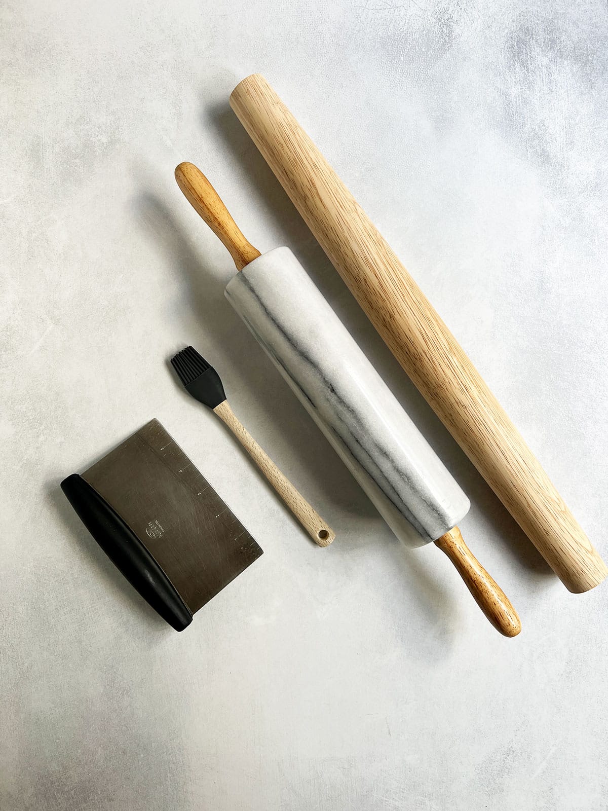 Tools that are helpful for making pie crust including a bench scraper, pastry brush, marble rolling pin and wooden rolling pin.
