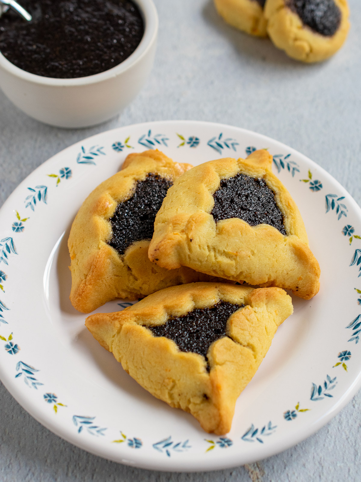 Close up of 3 poppy seed hamantaschen on a flowery plate with a bowl of filling in the background.
