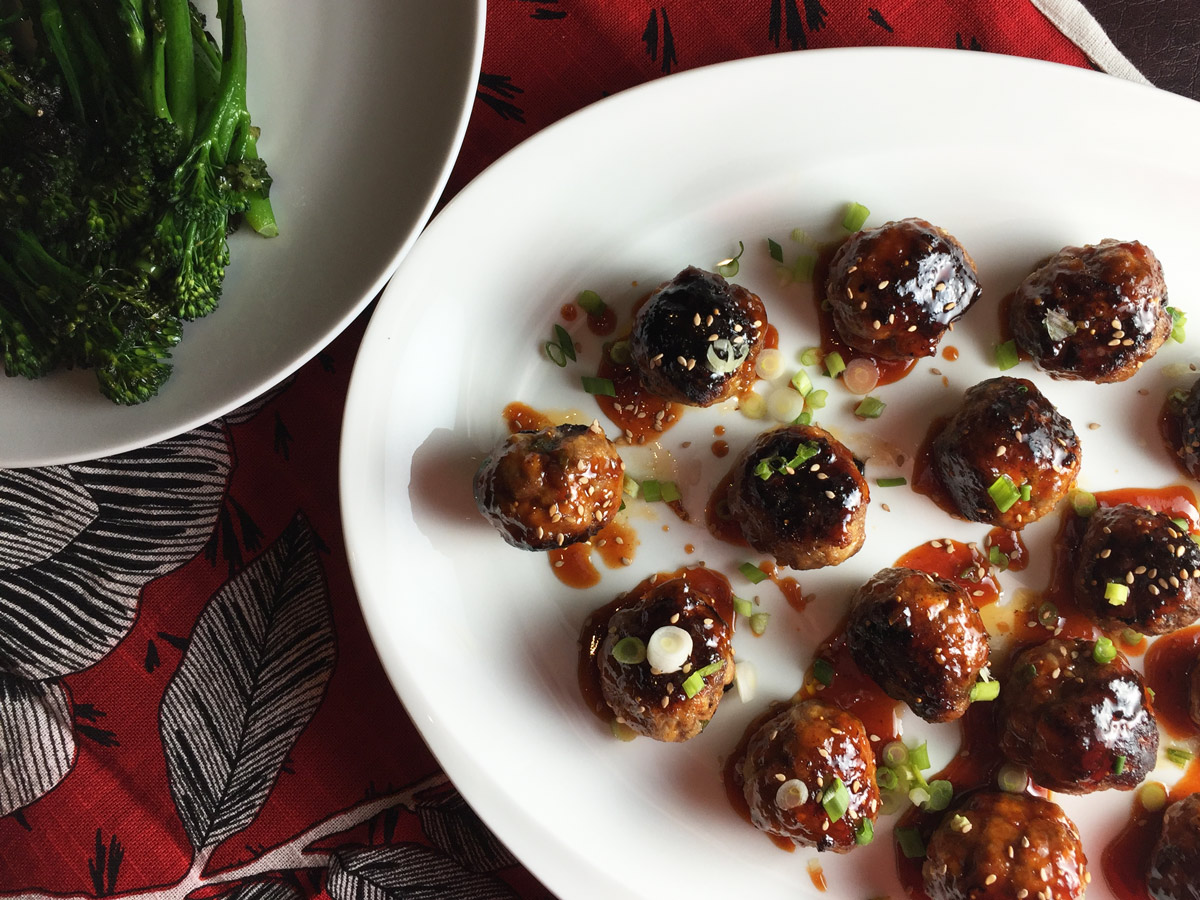 Glazed meatballs on a white plate with a plate of broccolini on the side.