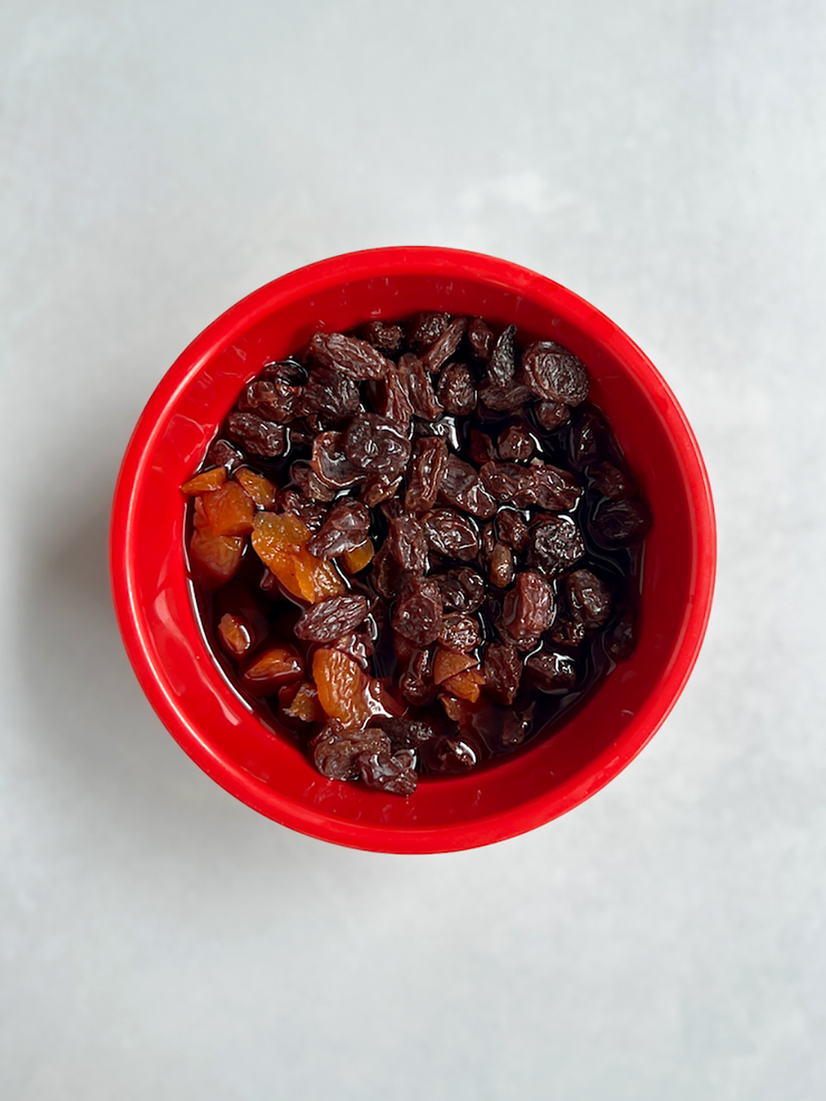 Raisins and apricots soaking in hot liquid in a red bowl.