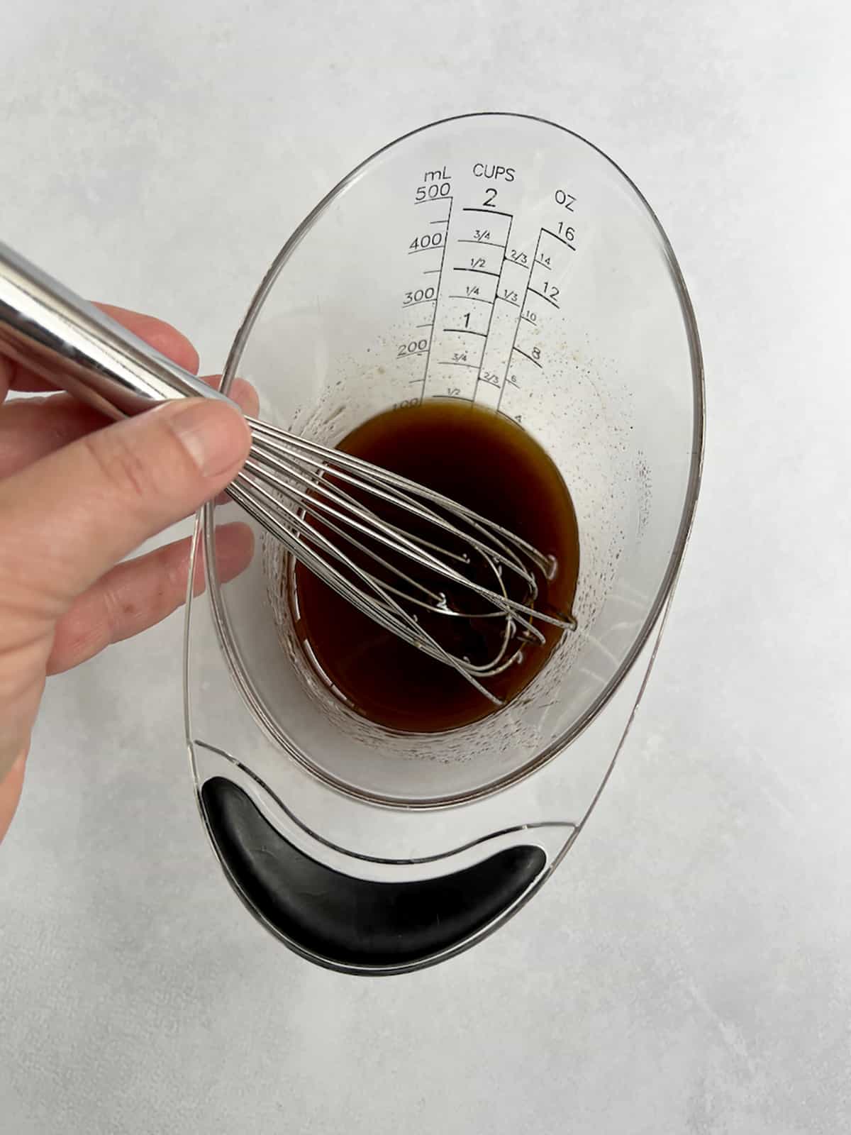 Liquid ingredients in a measuring cup with a hand holding a whisk.