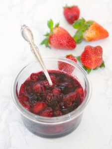 Fruit compote in a clear bowl with a silver spoon and strawberries in the background.