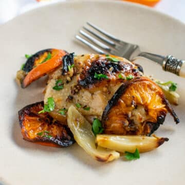 Chicken thigh with roasted clementines and fennel on a tan plate with a fork and a cut-open clementine in the background.