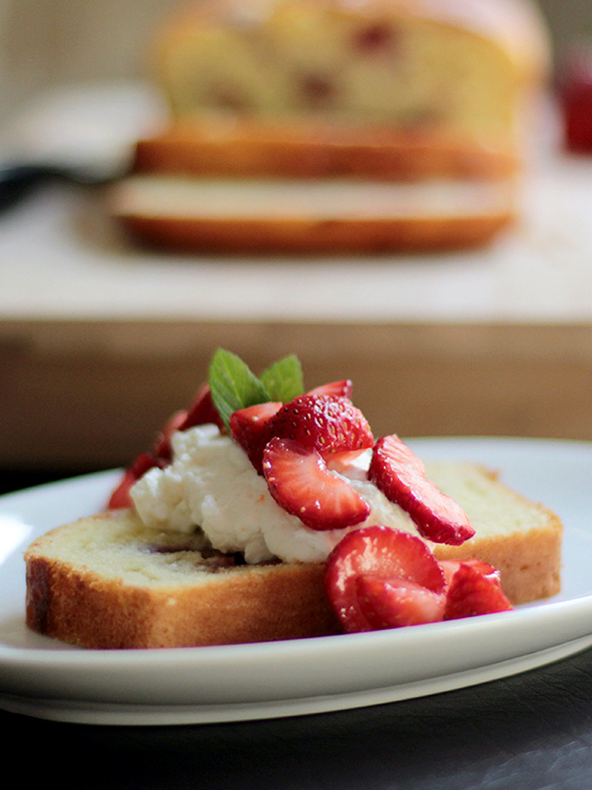 Slice of strawberry shortcake loaf on a white plate with the whole loaf in the background.