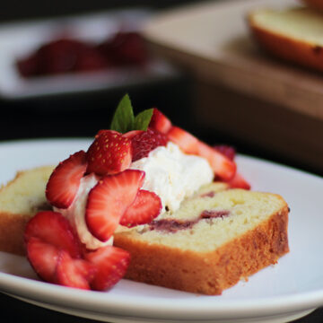 Strawberry shortcake loaf on a white plate with fresh strawberries and whipped cream on top with the rest of the loaf in the background.