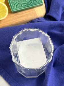 Square ice cube in a cocktail glass on a blue cloth napkin.