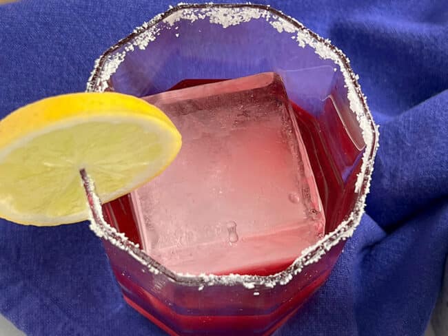 Straight down view of a cocktail glass with square ice cube inside and a lime on the rim plus salt and hibiscus margarita inside the glass, all on a blue napkin.
