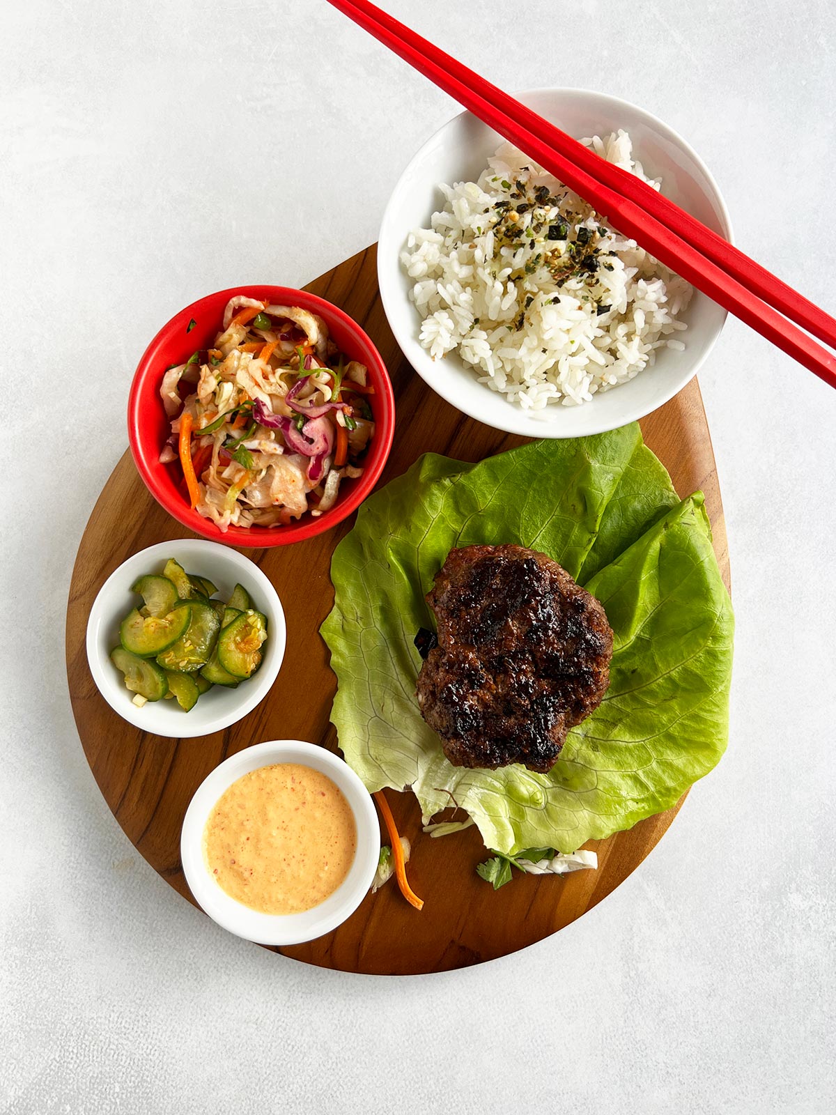 Burger on lettuce on a wooden board with kimchi sauce, Korean cucumbers, coleslaw and rice.