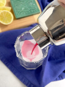Pouring hibiscus margarita over square ice in a salt-rimmed cocktail glass.