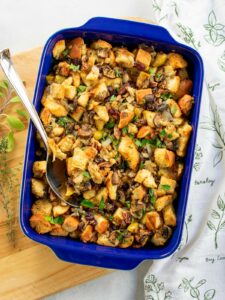 Mushroom challah stuffing in a blue casserole dish on a wooden board with a spoon in the stuffing.