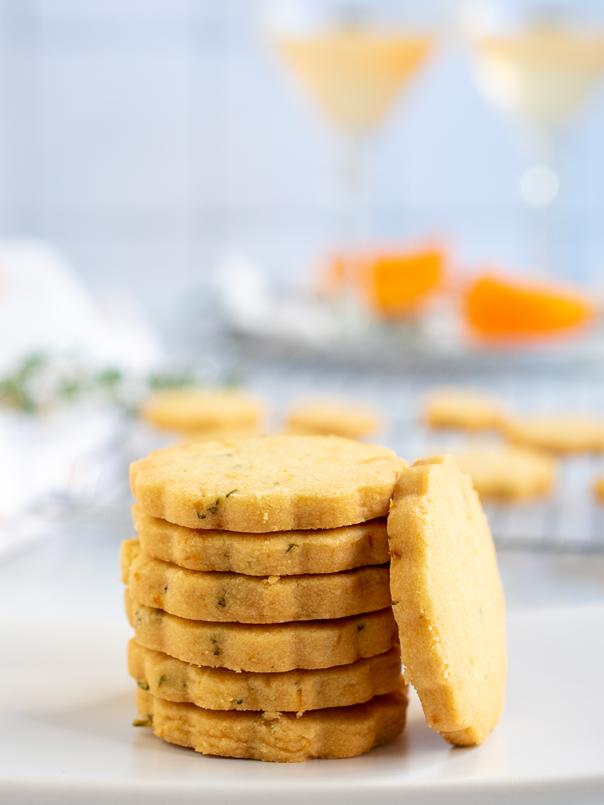 Straight on view of a stack of orange shortbread cookies on white plate with a view of two martini cocktails in the background.