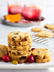 Cranberry orange shortbread cookies on a white plate with the cooling rack and two cocktails in the background.
