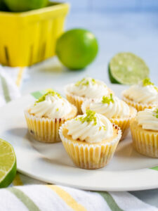 Several mini key lime pies on a striped white plate with limes in the background.