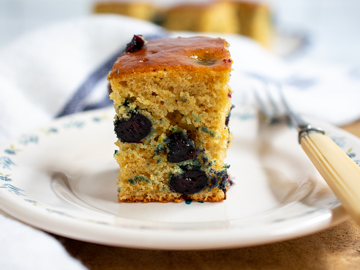 One slice of blueberry breakfast cake on a small off white plate with a fork on the side and more slices of cake in the background.