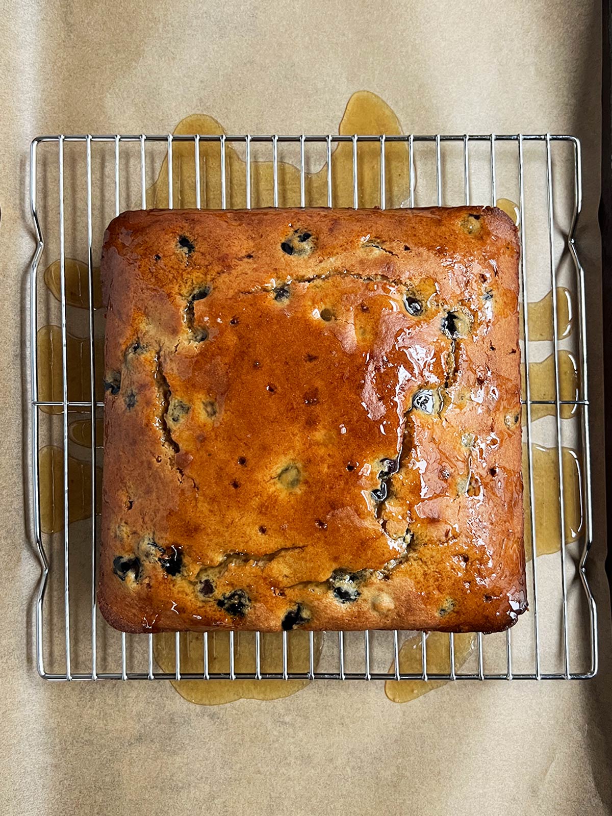 Olive oil blueberry cake on rack with glaze poured over and excess on the parchment paper below.