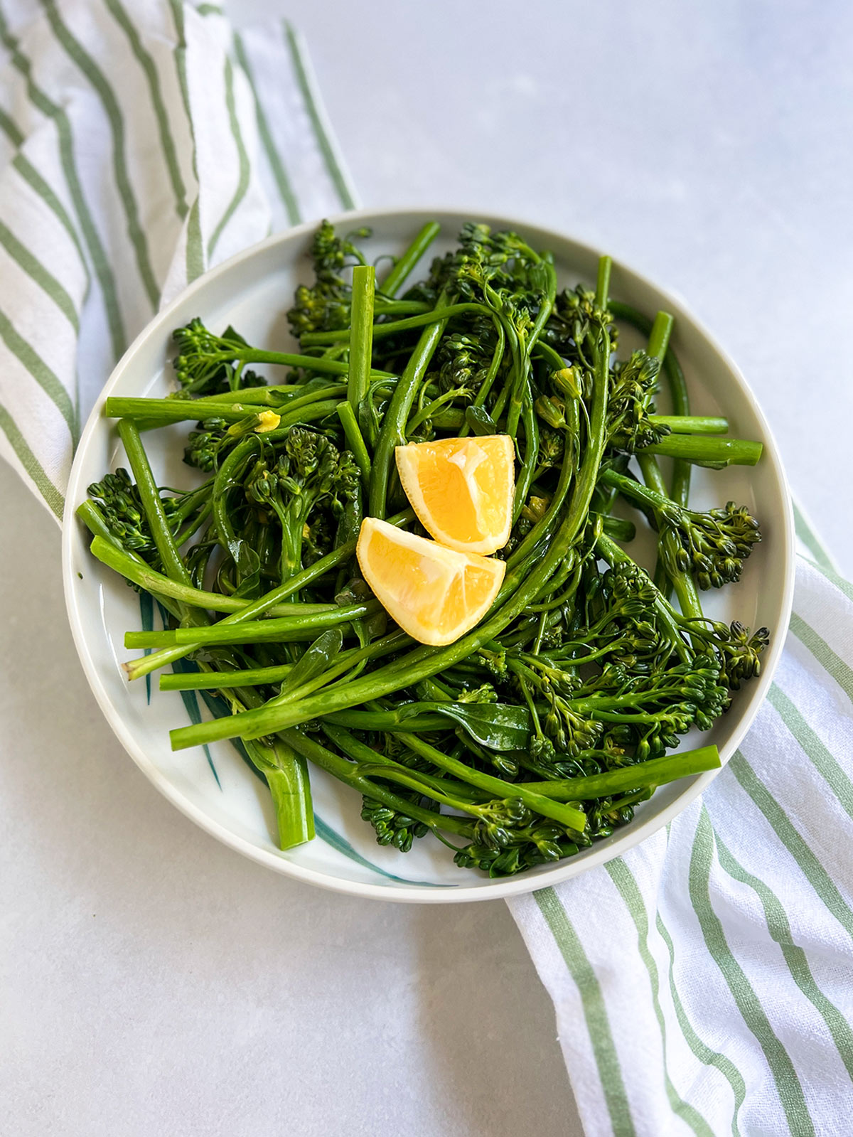 Broccolini on a white plate with a striped napkin underneath and w pieces of lemon on top.
