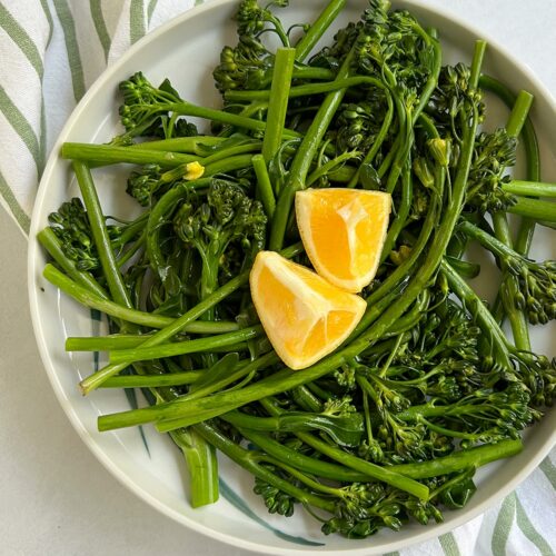 Braised broccolini on a plate with 2 pieces of lemon on top and a striped napkin underneath.