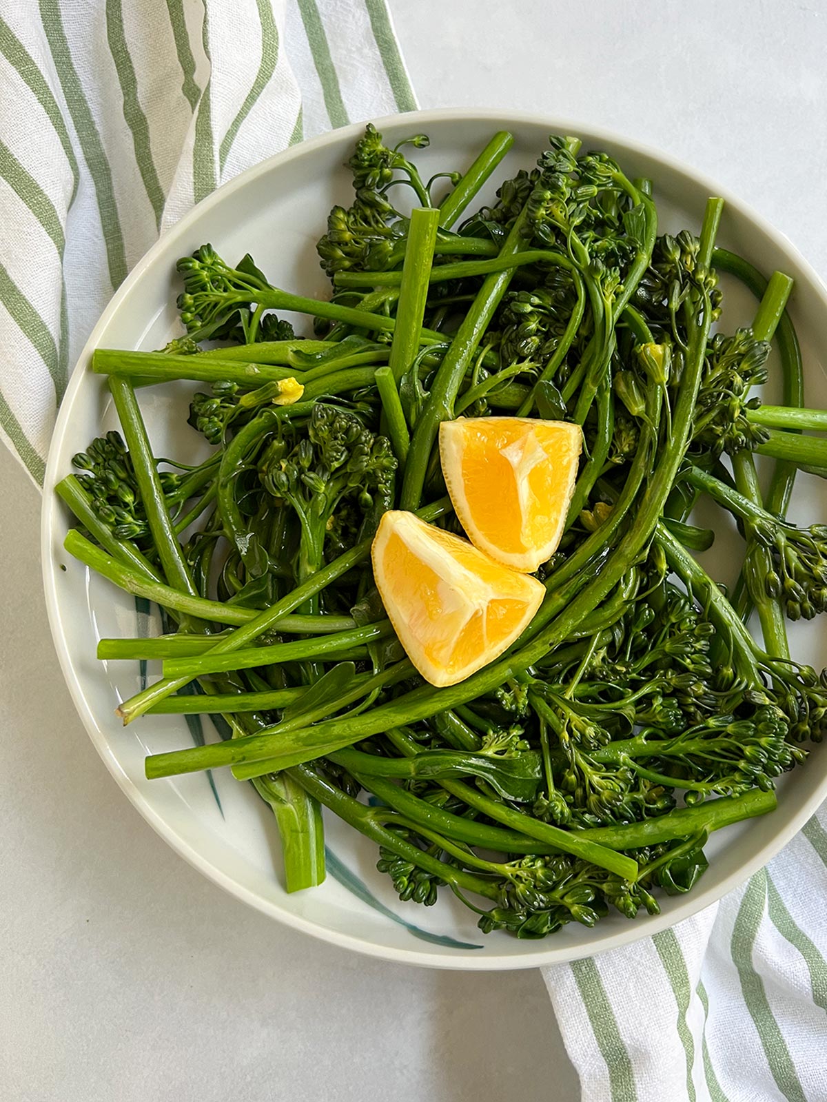 Braised broccolini on a plate with 2 pieces of lemon on top and a striped napkin underneath.