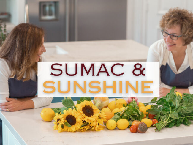 Sarene Wallace and Beth Lee leaning on a white kitchen counter with lots of fresh produce in front of them and words in the center that say Sumac & Sunshine.