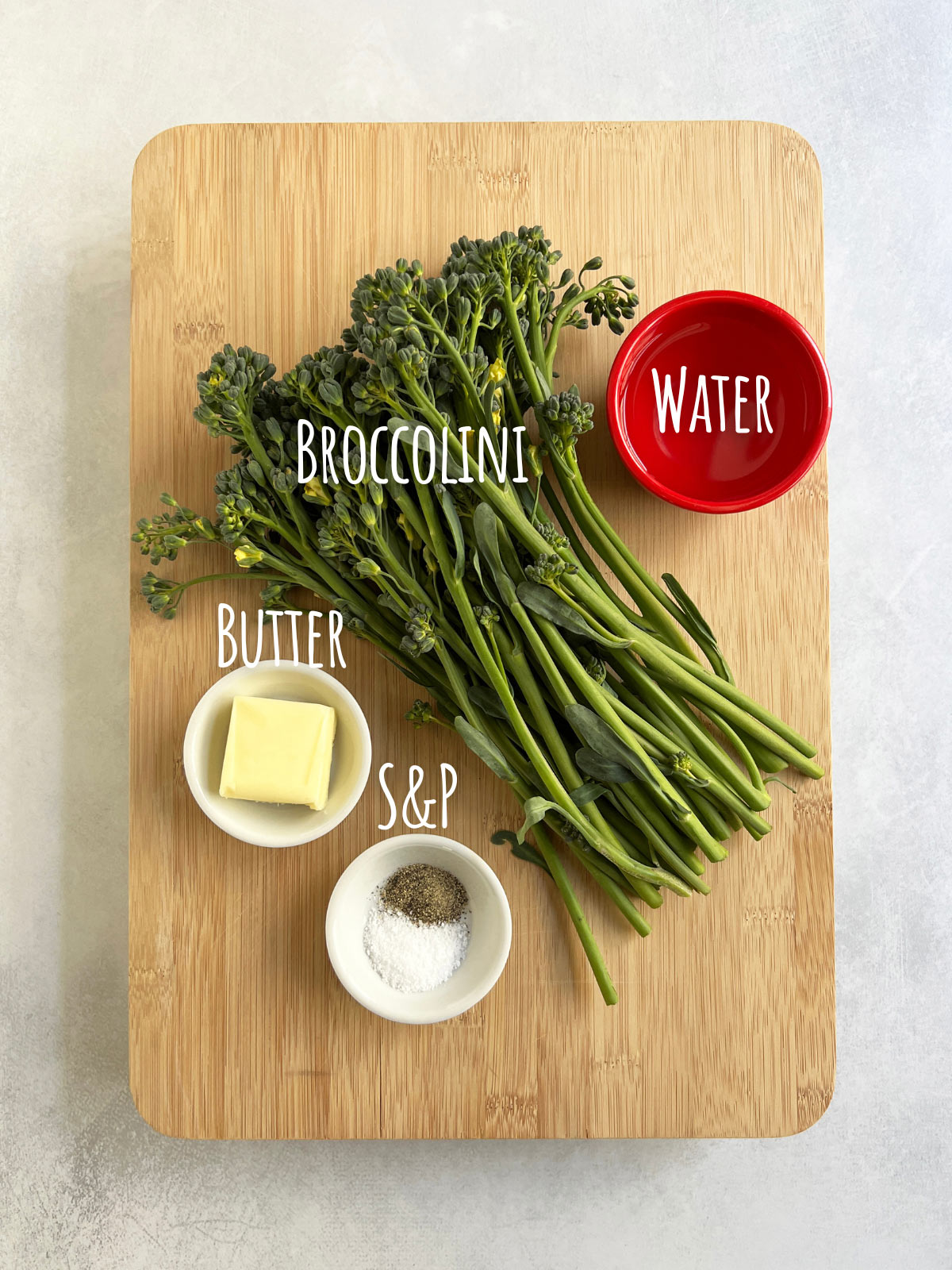 Ingredient shot for braised broccolini with vegetable, butter, salt, pepper, and water on a wooden cutting board.
