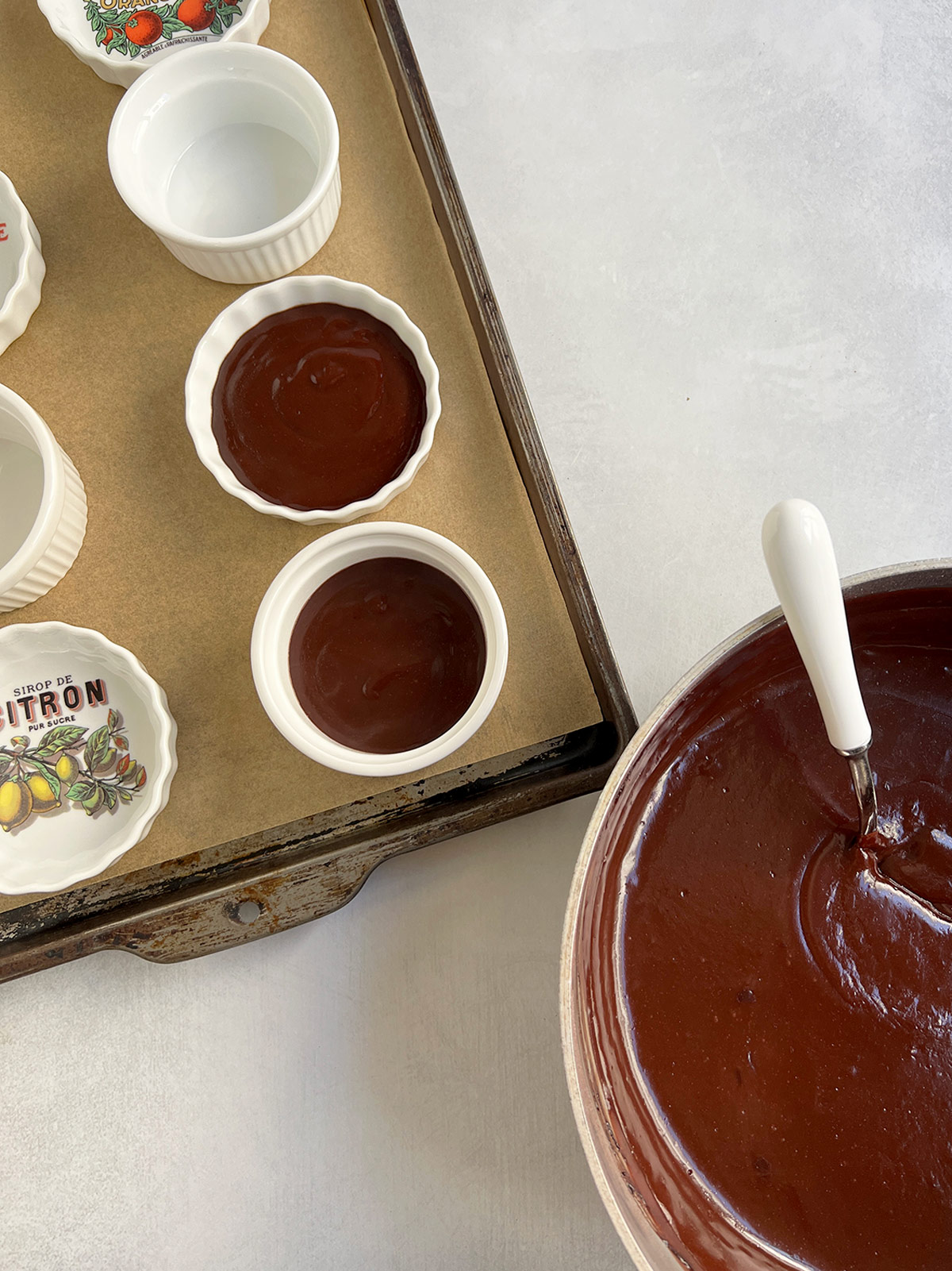 Filling bowls with dairy free chocolate pudding. Bowls are on a parchment lined sheet tray and pudding is still in the pot with a ladle.