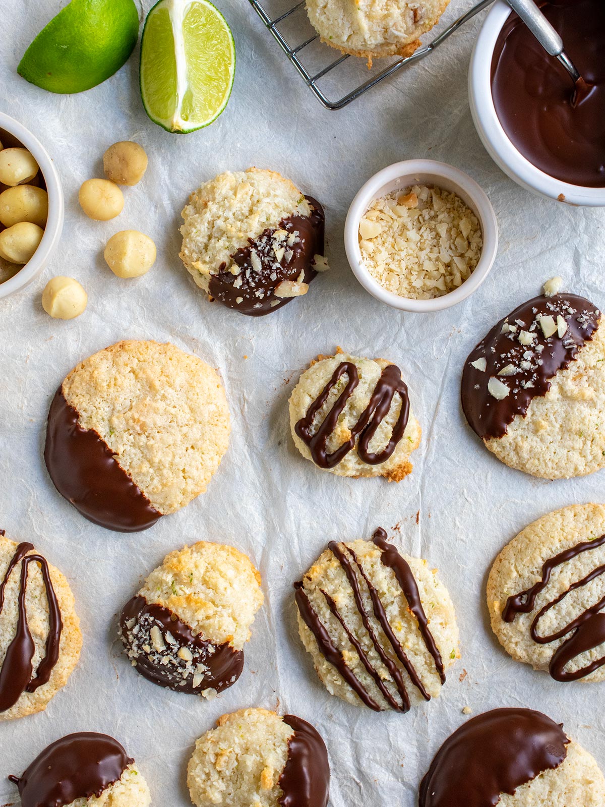 Flat lay of lots of dairy free macaroons both flat ones and puffy ones dipped and drizzled with a cooling rack and limes and a bowl of ganache plus macadamia nuts.
