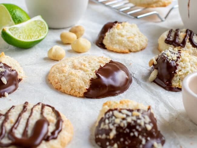 Close up of a chocolate dipped flat macaroon surrounded by other cookies and macadamia nuts and lime quarters.
