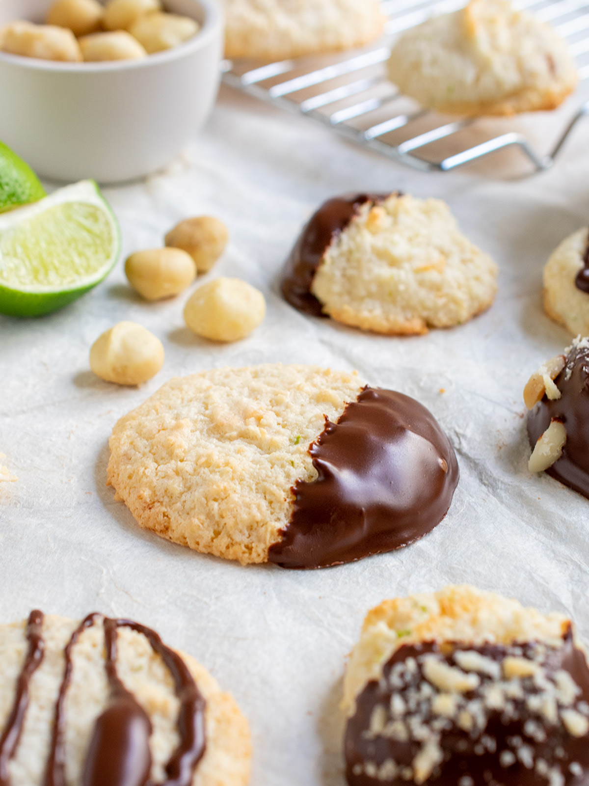 Angled view of a chocolate dipped macaroon with limes quarters and macadamia nuts and other cookies all around it.